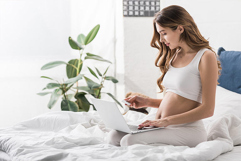 beautiful pregnant woman sitting on bed with laptop